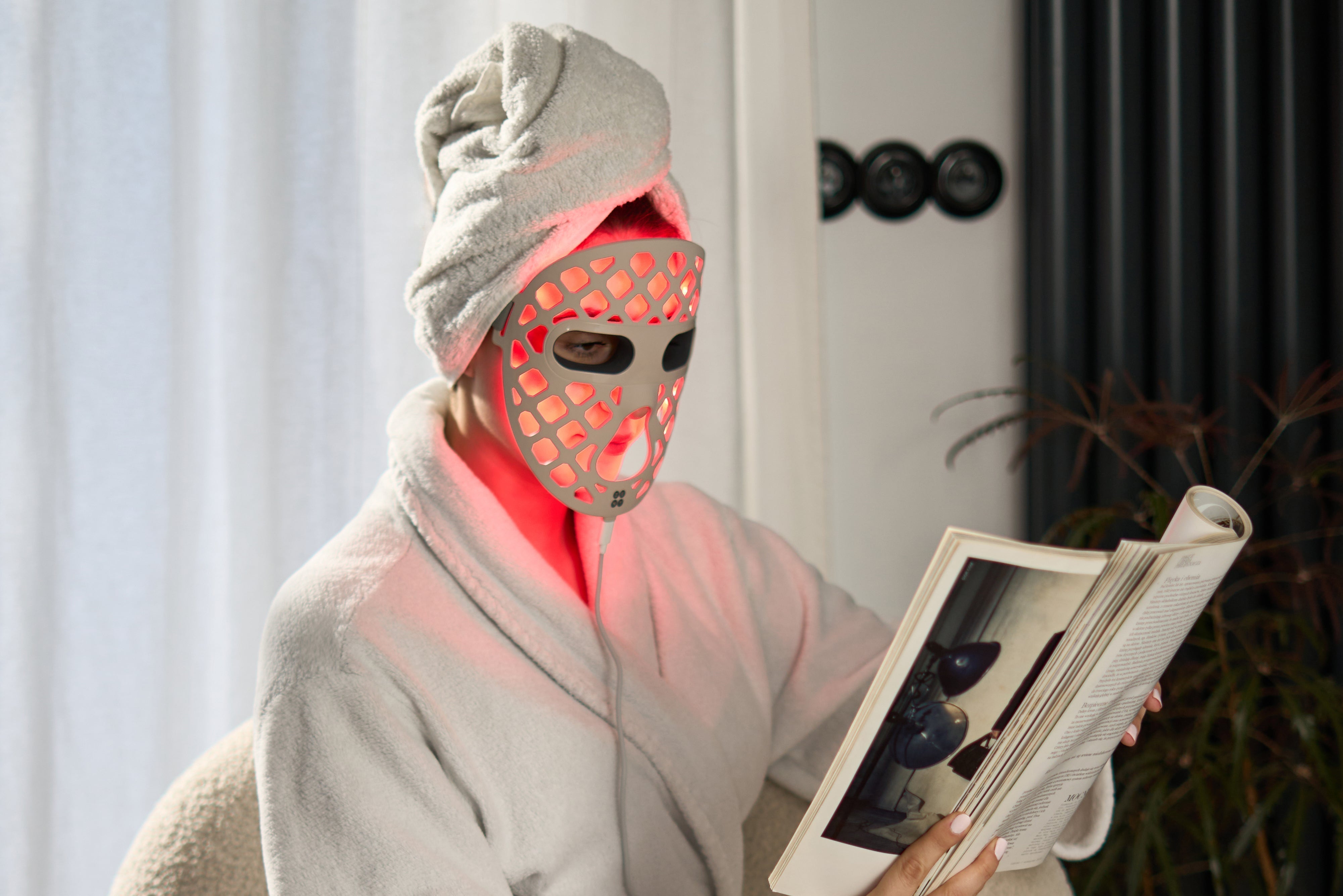 ZENT® DermaLight™ – Inteligentna maska LED do fototerapii 4w1 na twarzy kobiety, która czyta gazetę. Ubrana jest w szlafrok, a na głowie ma turban z ręcznika